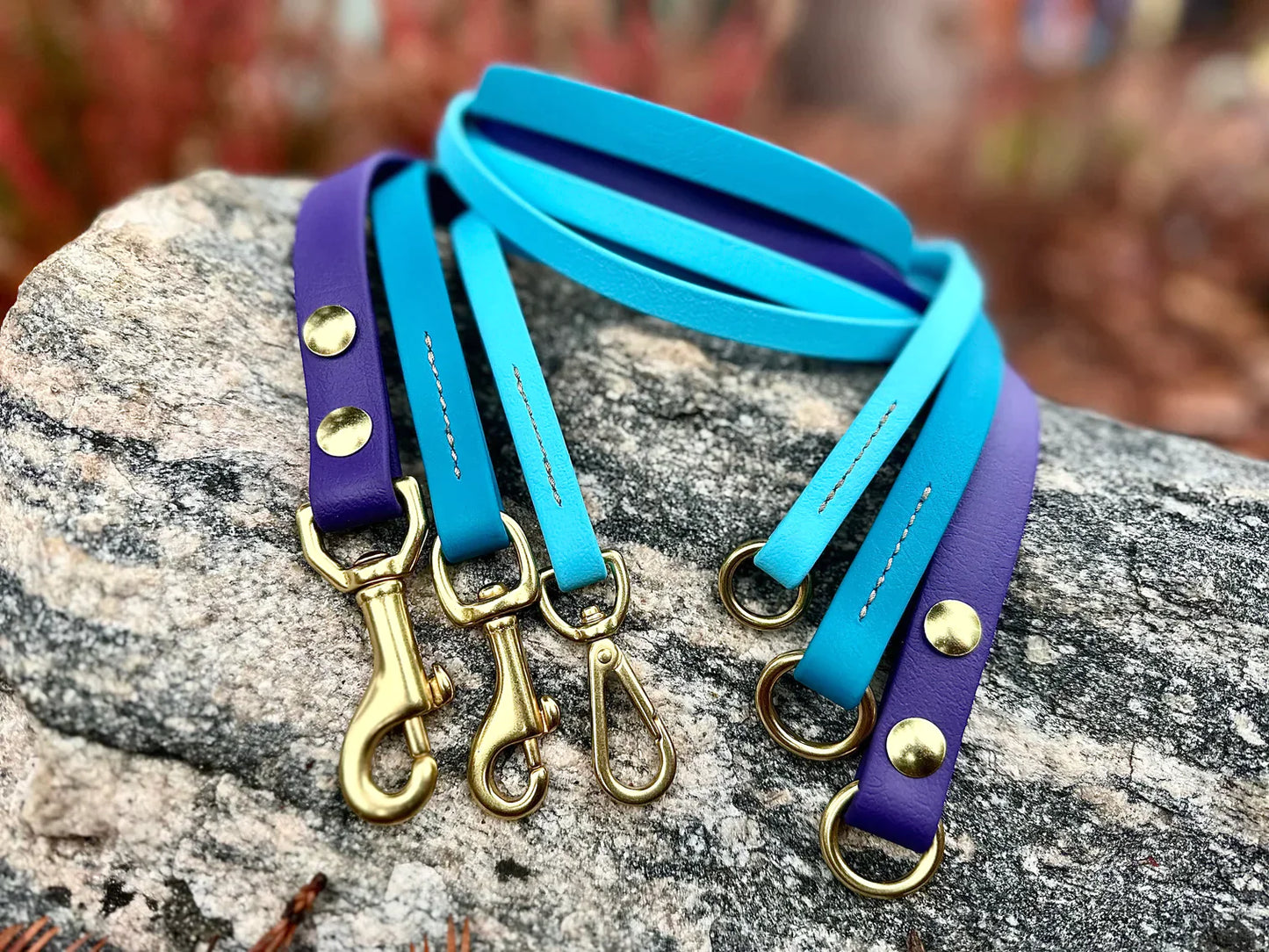 Three Your Whole Dog Trailblazing Tails: Long Line and Leash Extension (1/2 inch/Medium Dog) in blue and purple colors with gold metal clasps are laid on a textured rock surface.
