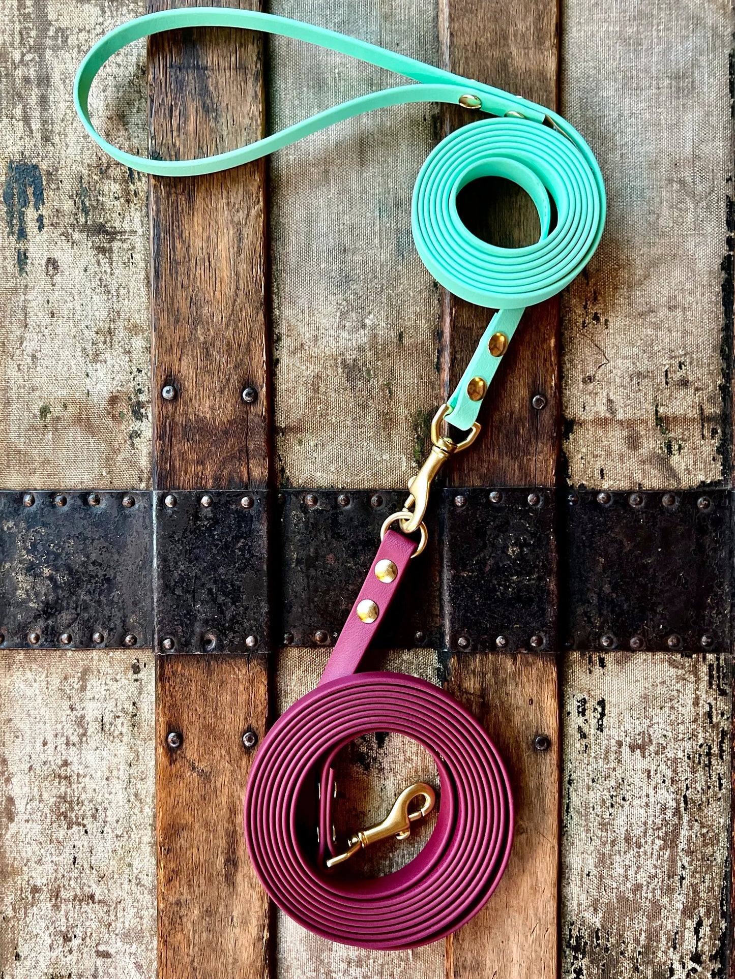 Two coiled, durable dog leashes lie on a weathered wooden surface with metal accents. One leash is mint green, and the other is burgundy, both featuring brass clasps. These are Trailblazing Tails: Long Line and Leash Extension (1/2 inch/Medium Dog) by Your Whole Dog.