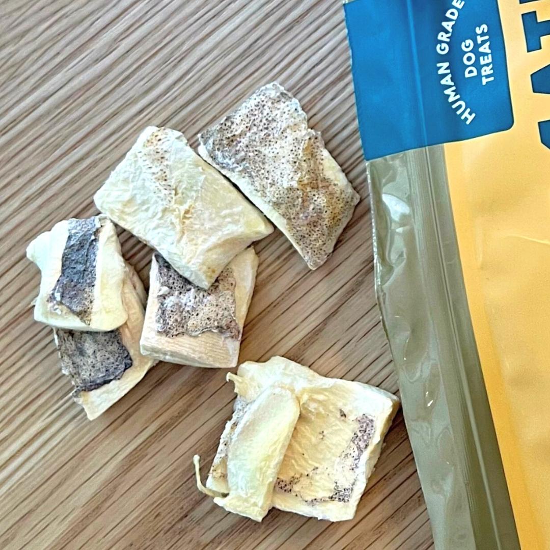 Scattered on the wooden surface are several dog treats beside an open yellow and blue package labeled "Gourmate Pet Treat Co. Wild Caught Calamari," described as human-grade.
