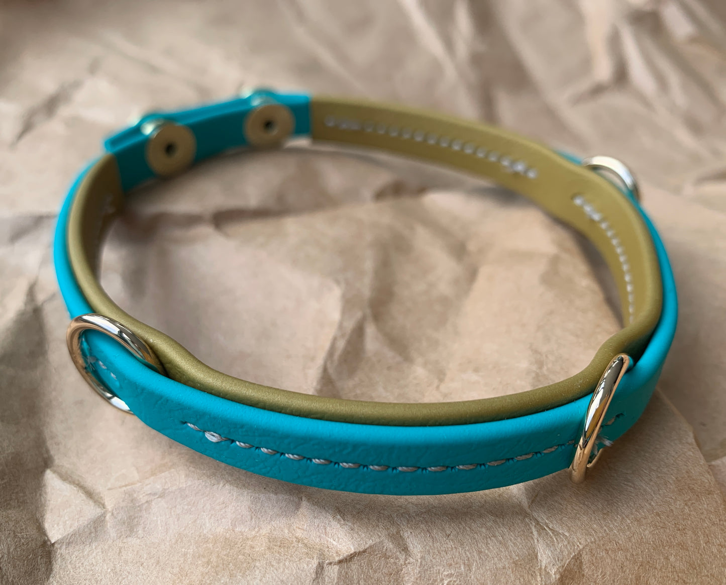 A close-up of a durable blue and green handcrafted Trailblazing Tails: The Leash Organiser by Your Whole Dog with two metal D-rings and silver snap buttons, lying on a crumpled brown paper surface.