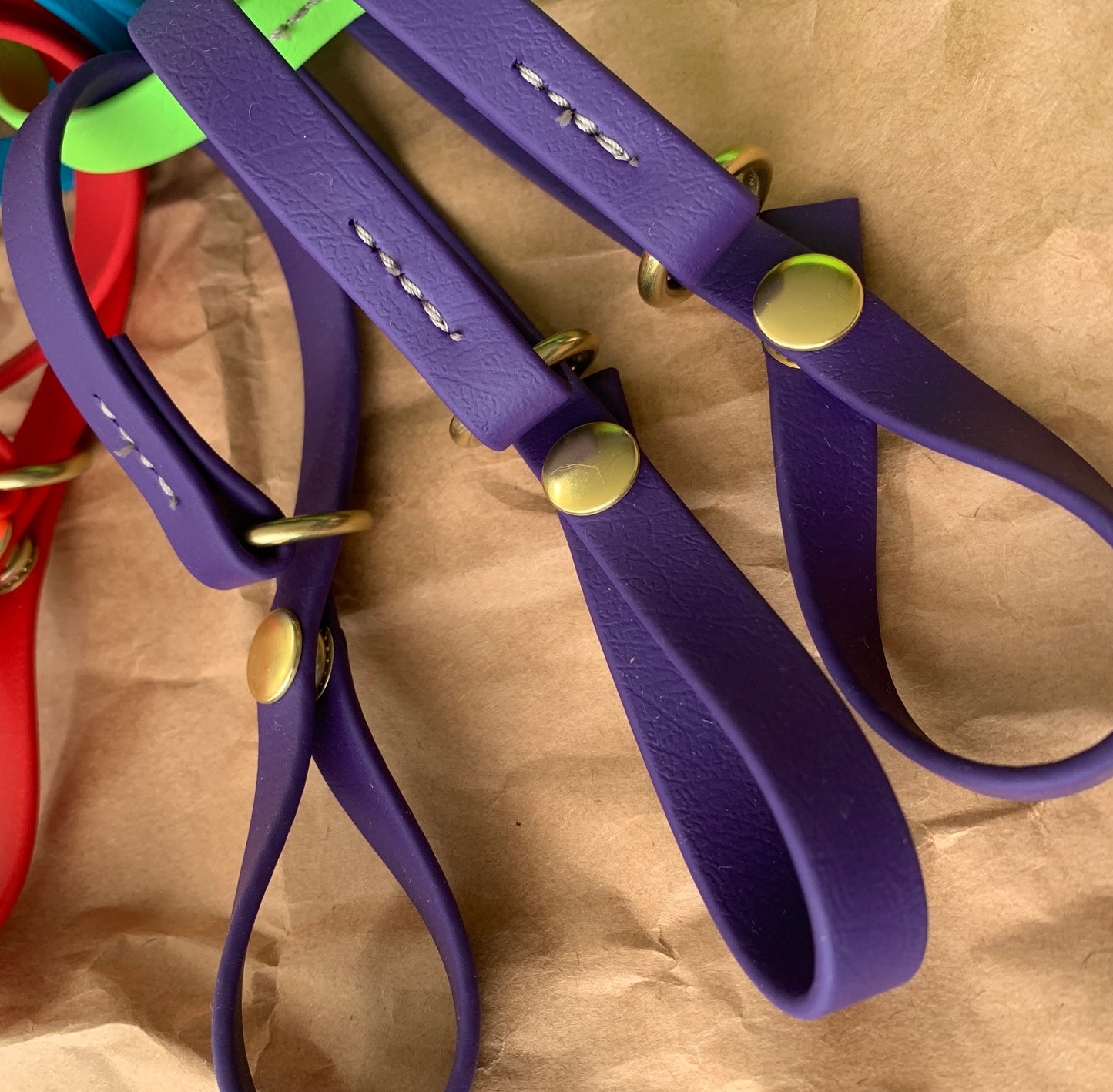 A close-up of purple leather straps with brass buttons and metal rings, set atop a brown paper surface, with parts of red and green straps visible in the background. Ideal for those needing to cinch your line effectively, the Trailblazing Tails: The Long Line Holder from Your Whole Dog meets all your needs.
