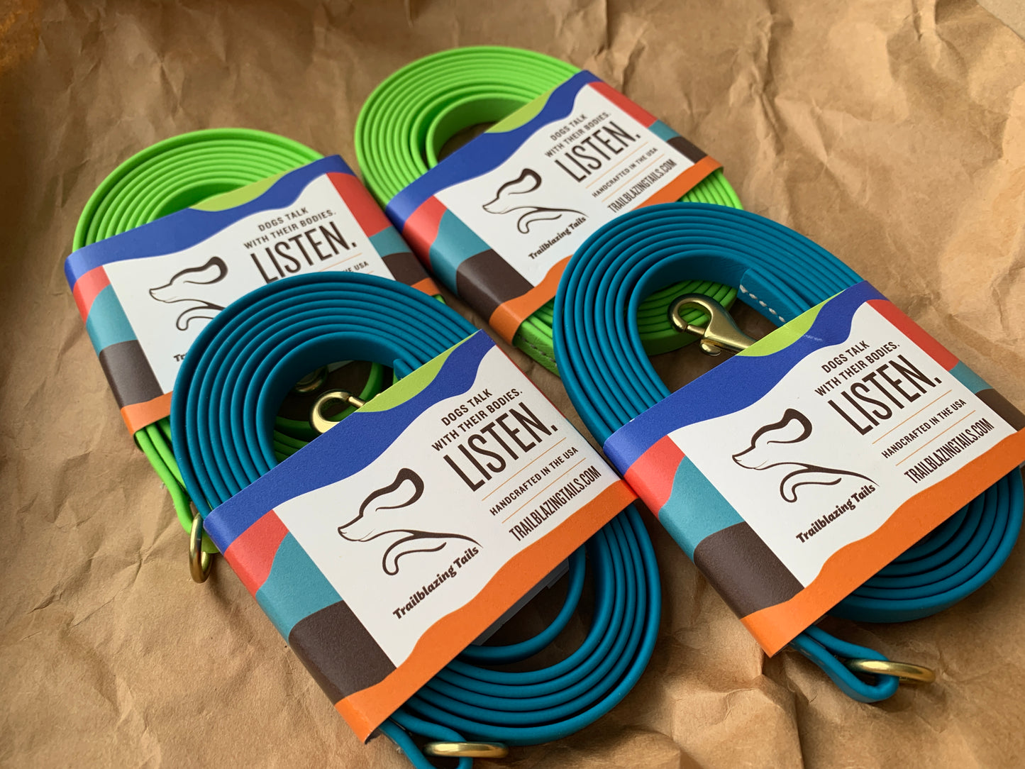 Four coiled, durable Trailblazing Tails: Long Line and Leash Extension (1/2 inch/Medium Dog) leashes in green and blue, each with a colorful label featuring a dog silhouette and the text "Listen! Dress your dog with pride." They are placed on crumpled brown paper.