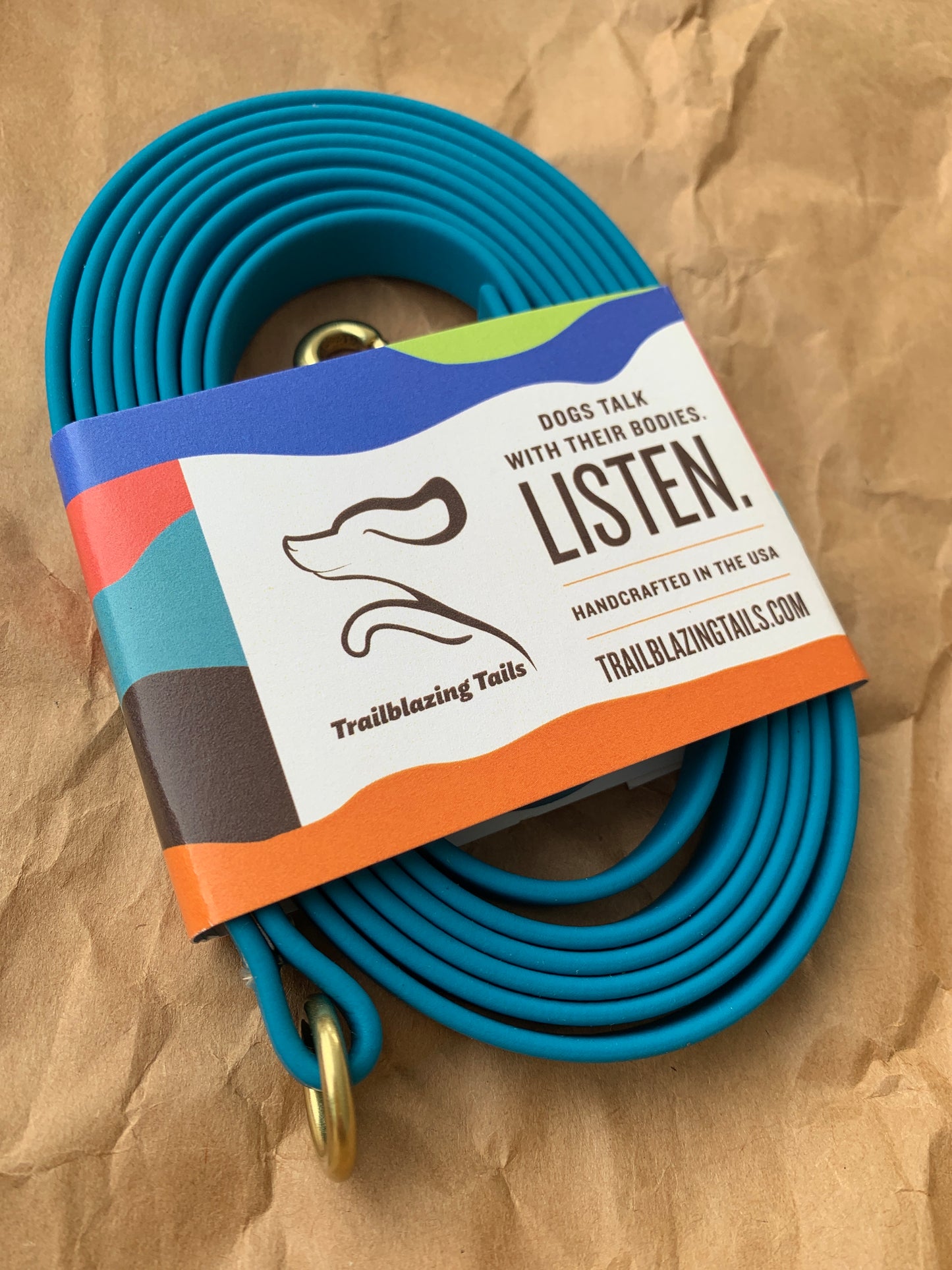 A coiled blue, durable dog leash with a brass clasp, wrapped in a label reading, "Dogs talk with their bodies. Listen. Handcrafted in the USA." and branded "Trailblazing Tails: Long Line and Leash Extension (1/2 inch/Medium Dog)" by Your Whole Dog.