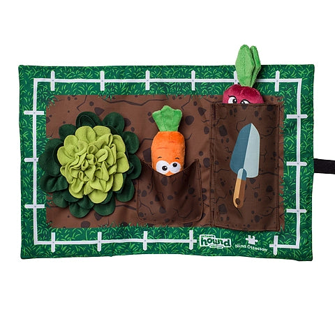The Nina Ottosson: Interactive Snuffle Activity Puzzle Mat by Your Whole Dog is a garden-themed dog toy with pockets holding a stuffed carrot, a stuffed radish, and a soft trowel. It features an attached fabric plant and a background grid pattern, making it perfect for hiding treats to provide mental exercise.