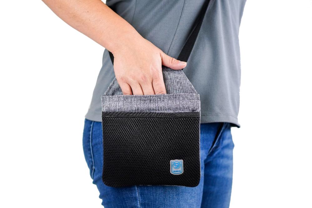 A person in a gray shirt and jeans holds the Blue-9 INSPIRE Training Treat Pouch, a small black and gray shoulder bag with a blue logo, ideal for carrying training treats.