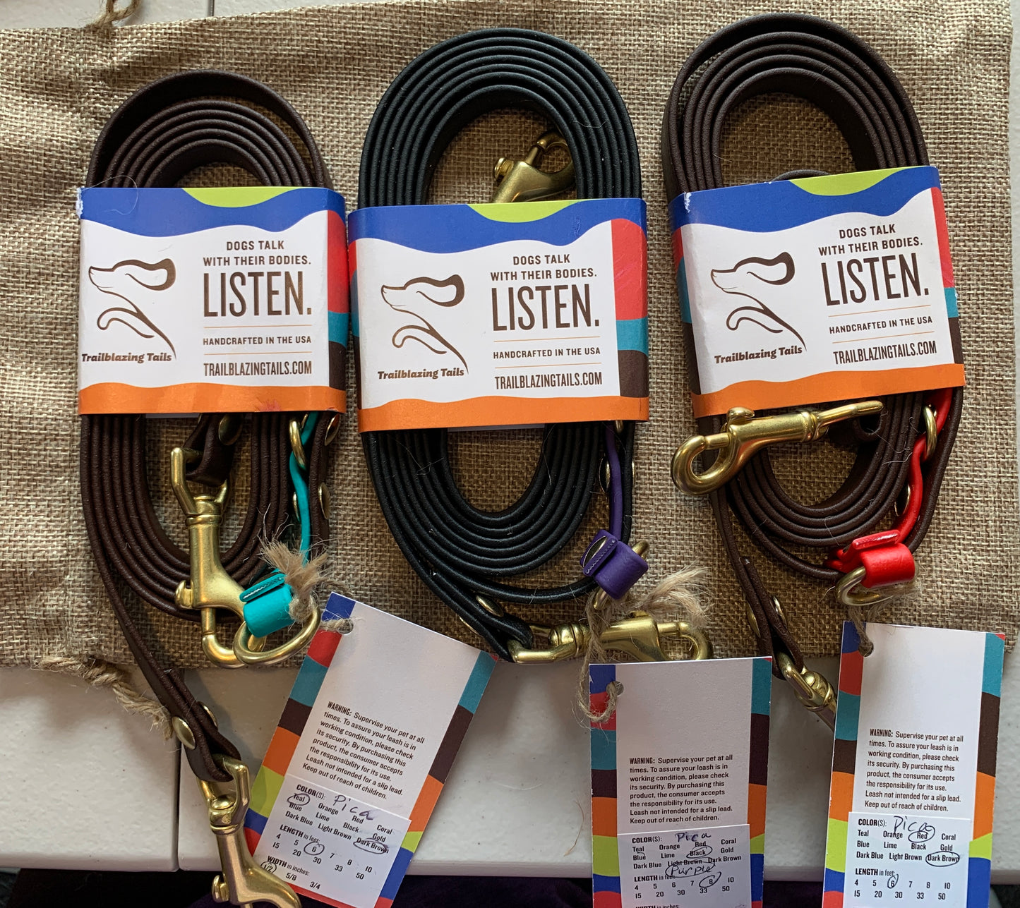 Three neatly coiled leashes with tags attached are displayed. Each leash features a brass clip and a label that reads "Dogs talk with their bodies. Listen." Experience hands-free innovation with Your Whole Dog's Trailblazing Tails: The Pica, designed for the modern pet owner.