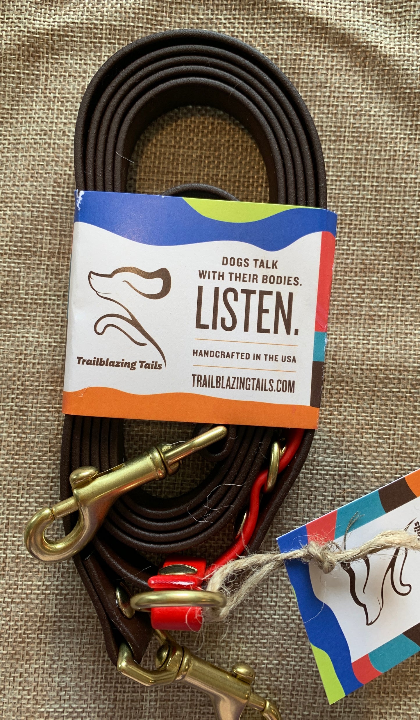 A coiled brown dog leash with brass hardware is displayed. Attached is a label reading, "Dogs talk with their bodies. Listen. Handcrafted in the USA" and the brand name "Your Whole Dog." Experience hands-free innovation with this beautifully crafted piece: Trailblazing Tails: The Pica.