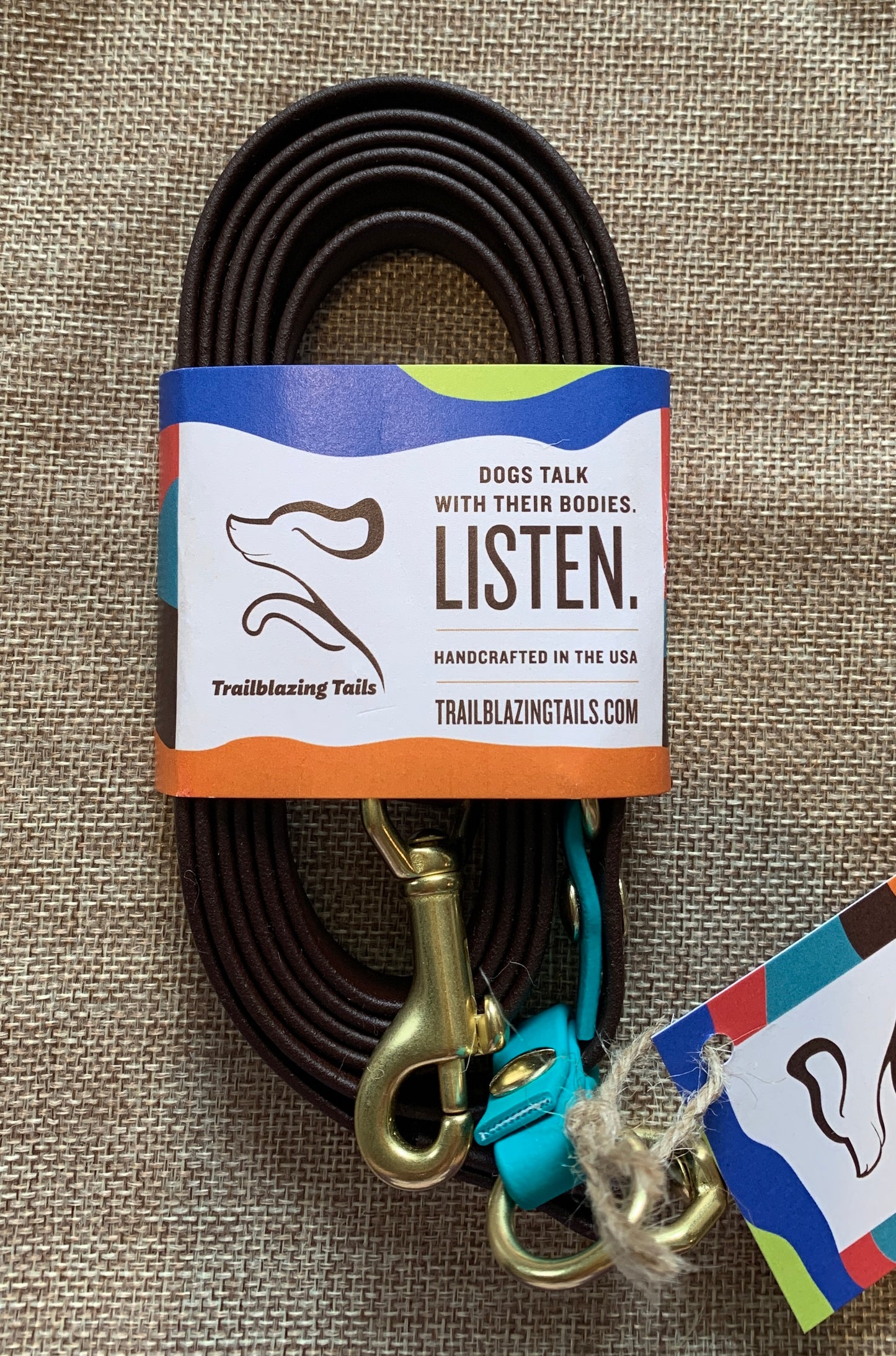 A coiled brown dog leash with brass hardware and a blue clip. The Pica, by Your Whole Dog, is wrapped with branding that reads, "Dogs talk with their bodies. Listen." Handcrafted in the USA, this dog leash brings hands-free innovation to your daily walks.