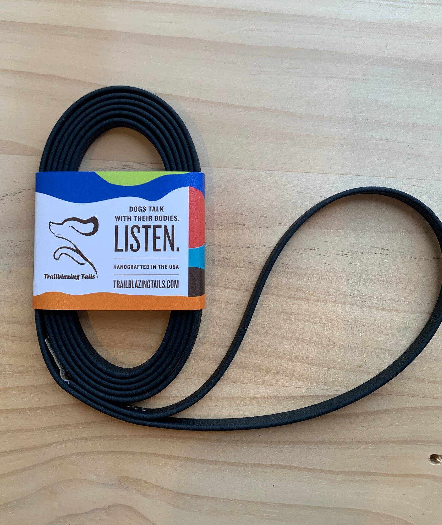 A coiled black dog leash with a colorful label showing a dog's face profile and text: "Dogs talk with their bodies. Listen." The label also mentions the website trailblazingtails.com and its Slide Tab Handle design. This ultra-lightweight leash is handcrafted in the USA by Your Whole Dog and is named Trailblazing Tails: The Little Dog.