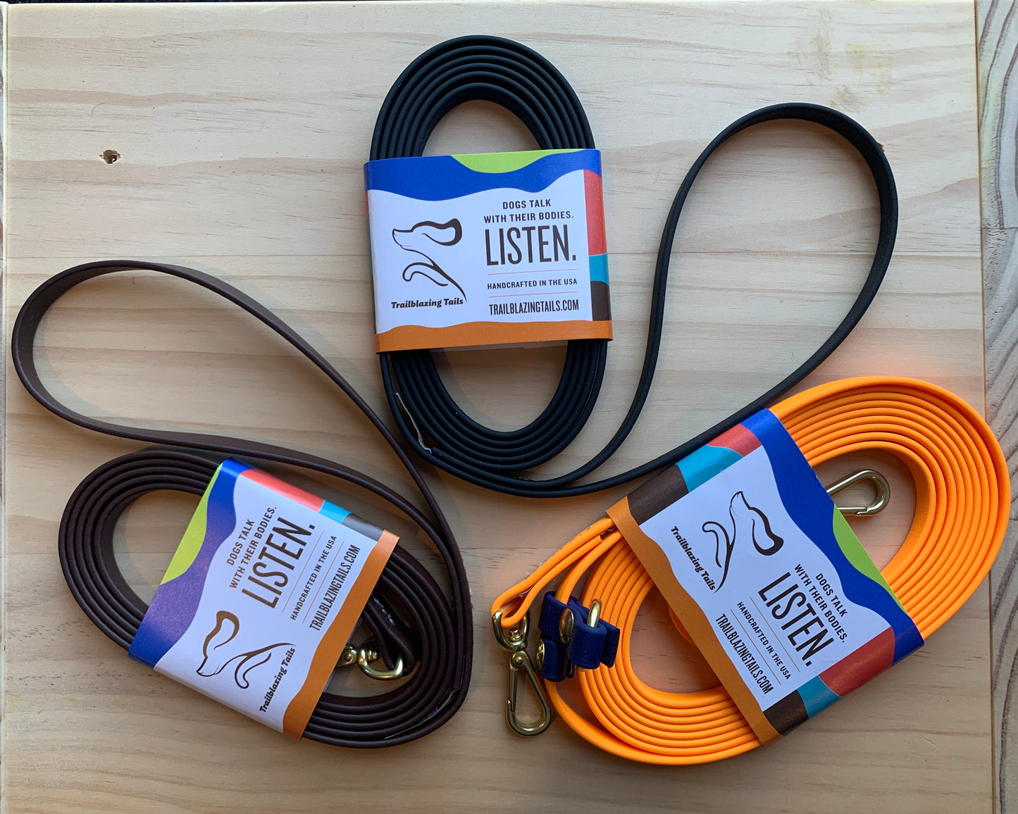 Three ultra-lightweight, coiled Trailblazing Tails: The Little Dog leashes of different colors (black, brown, and orange) from Your Whole Dog are displayed on a wooden surface. Each leash, designed to be odour-proof, features a label with the text, “Dogs talk with their noses. Listen.”