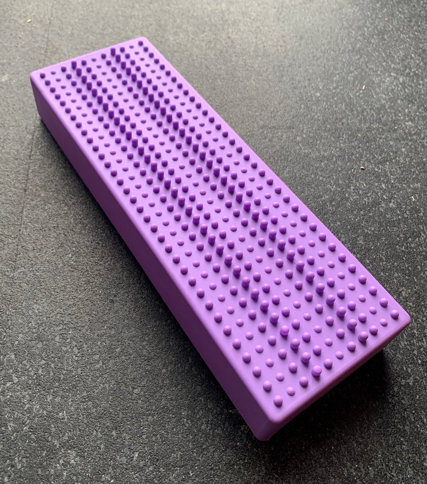 The purple rectangular spiky massage mat fits seamlessly on a dark surface, designed with Flexiness StackingBar compatibility for enhanced comfort.