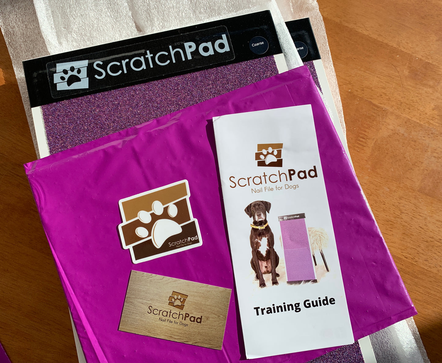 The ScratchPad for Dogs Nail Scratch Boards, Fear Free® endorsed, come with a training guide, sticker, and packaging beautifully presented on a wooden surface.
