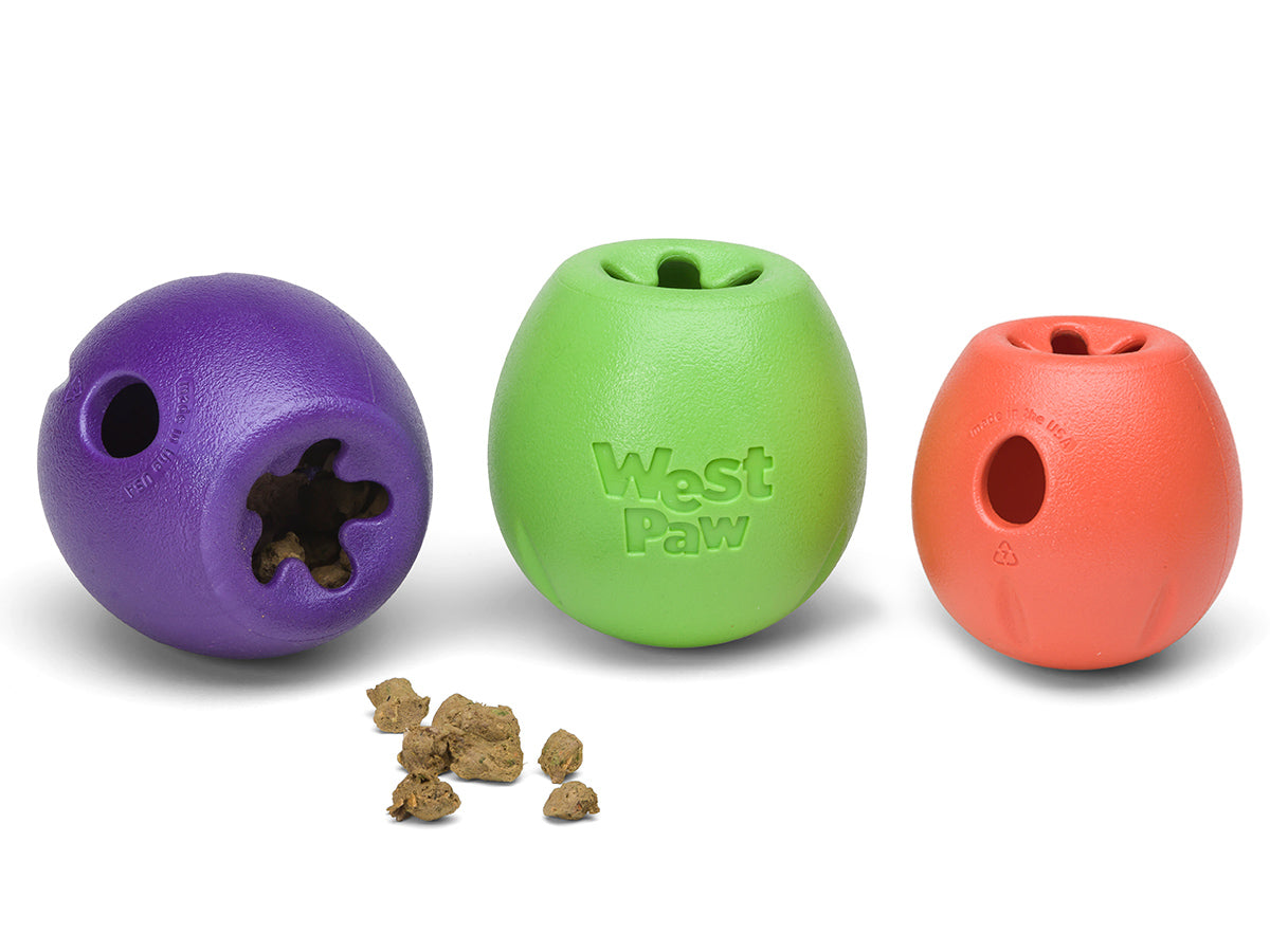 Three colorful West Paw Rumbl treat-dispensing puzzle toys sit side-by-side, with scattered dog treats in front—perfect as durable West Paw dog toys to keep your pup engaged.