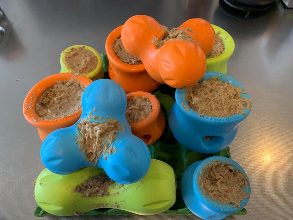 West Paw Tux treat toys, designed for tough chewers, are on a table filled with a beige, mushy mixture. Available in blue, orange, and green colors, these toys are also conveniently dishwasher safe for easy cleaning.
