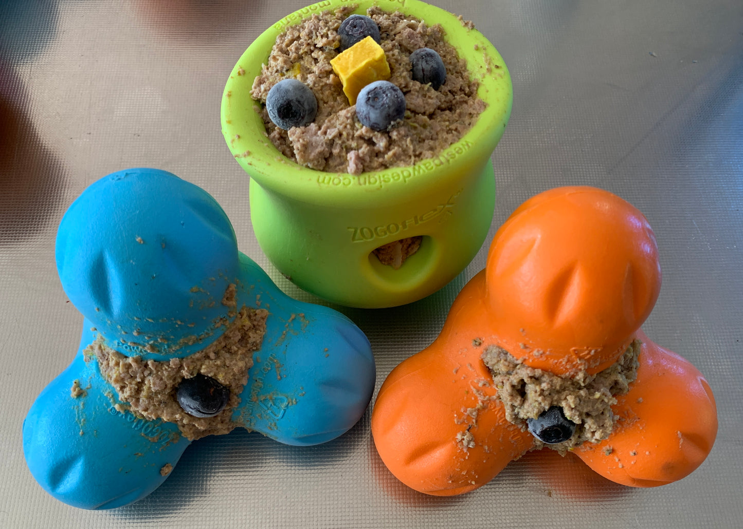 The West Paw Tux by Your Whole Dog includes three colorful, interactive toys ideal for tough chewers, filled with kibble and blueberries. The green treat toy has a yellow cube on top. All are dishwasher safe for easy cleaning.