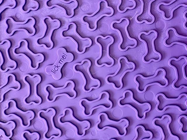 Explore the Soda Pup EMAT Enrichment Licking Mat, a purple textured silicone mat featuring bone shapes for dog foraging. One bone humorously says "lick me," making it ideal for stimulating and engaging your dog's mind and senses.