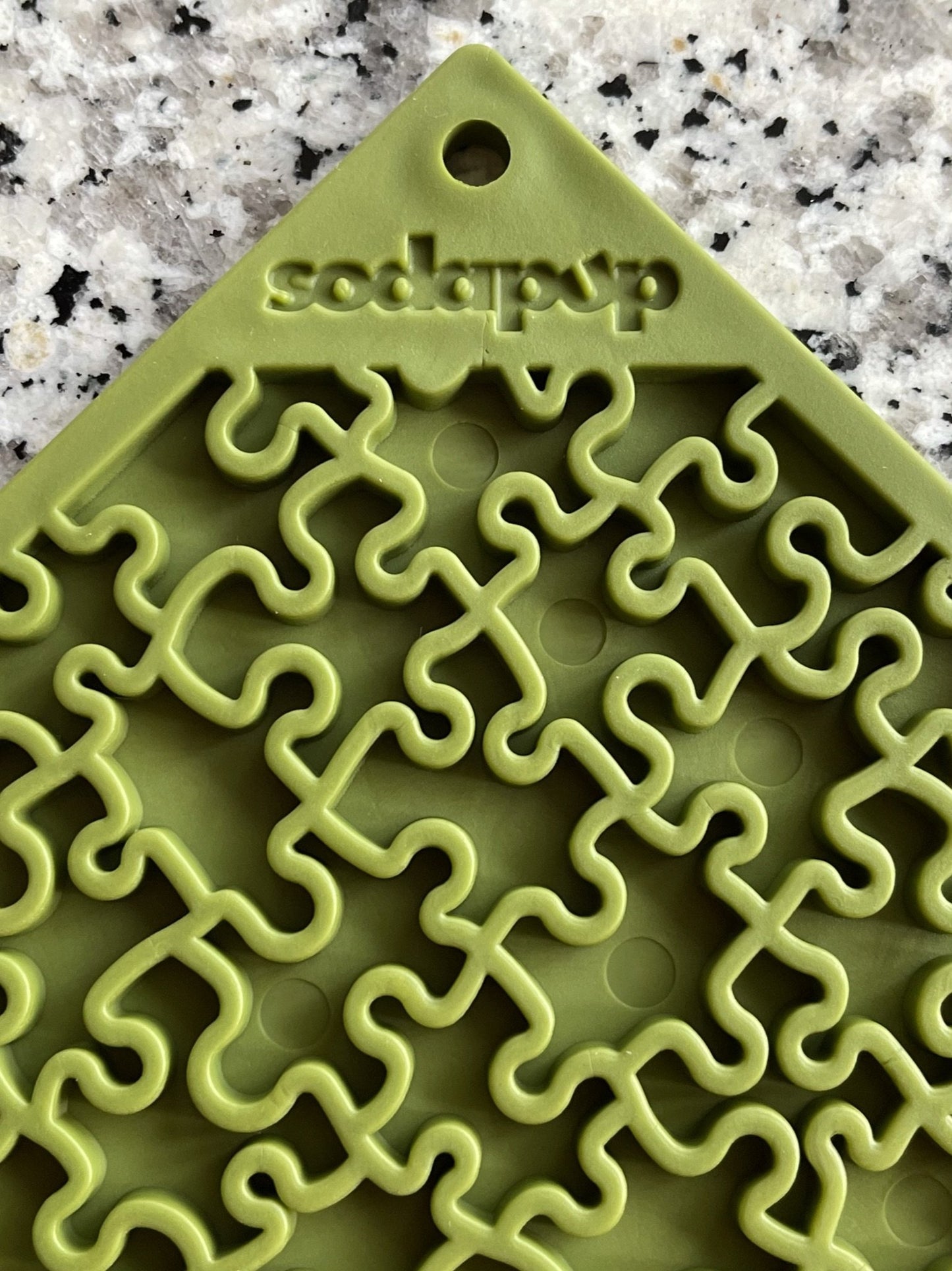 The "CLEARANCE: Soda Pup EMAT ENRICHMENT LICKING MAT" is a green silicone mat with a puzzle-piece design, embossed with "sodapup," ideal for dog foraging and engaging in playful licking toy activities. Displayed on a speckled countertop, it enriches your pup's experience.