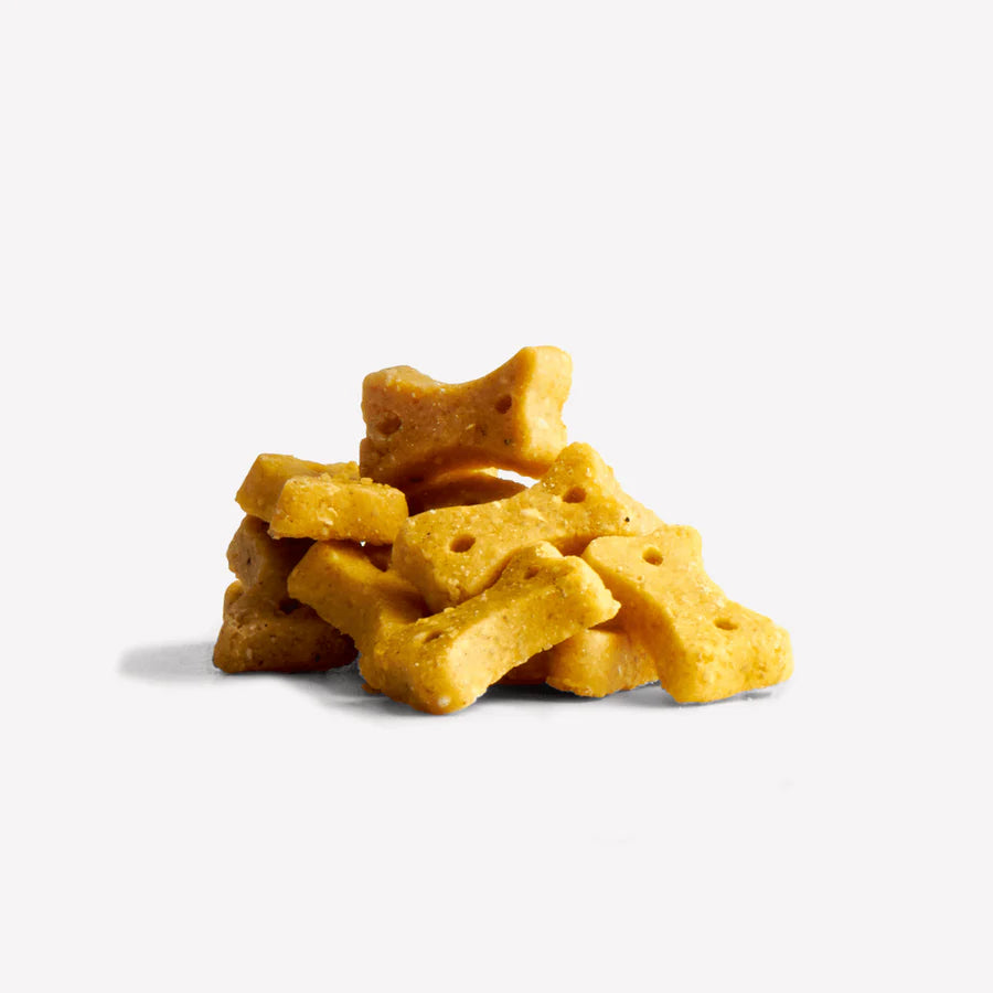 A small pile of Beco Pets Peanut Dog Treats with Coconut & Turmeric, bone-shaped and yellowish, rests on a white background.