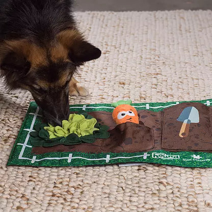 A dog is sniffing at the Nina Ottosson Interactive Snuffle Activity Puzzle Mat by Your Whole Dog, designed to look like a garden with a green patch, plush carrot, and small shovel on a textured surface—perfect for treat hiding and mental exercise.