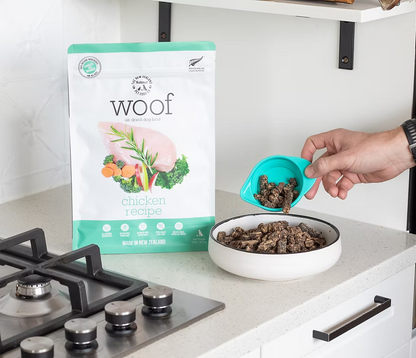 A hand pours WOOF Air Dried Chicken Dog Food by WOOF - The NZ Natural Petfood Co into a bowl, with a bag of premium New Zealand dog food on the kitchen counter nearby.