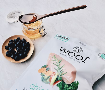 A jar of honey with a wooden spoon, a small bowl of blueberries, and a pack of WOOF Freeze Dried Chicken Dog Food by WOOF - The NZ Natural Petfood Co are arranged on a light surface.