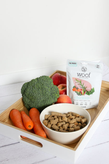 A tray with broccoli, carrots, apples, a bowl of natural dry dog food, and a packet of WOOF Freeze Dried Chicken Dog Food by WOOF - The NZ Natural Petfood Co is displayed on a white surface.