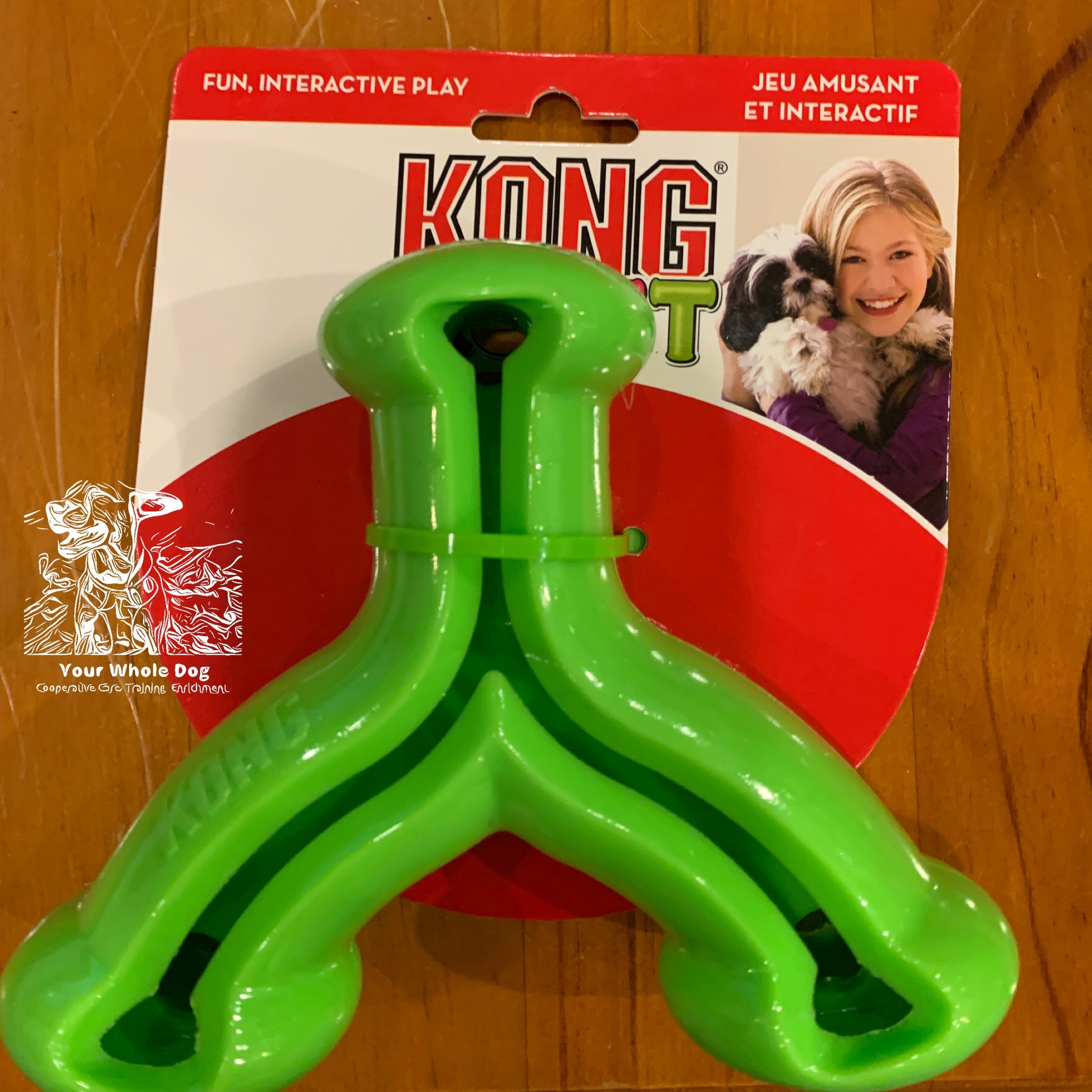 A green Your Whole Dog KONG Quest Wishbone dog toy in packaging featuring a picture of a person holding a dog on the top right corner. The text on the packaging reads "Fun, Interactive Play / Jeu Amusant et Interactif." This treat-dispensing toy encourages natural foraging behaviors.