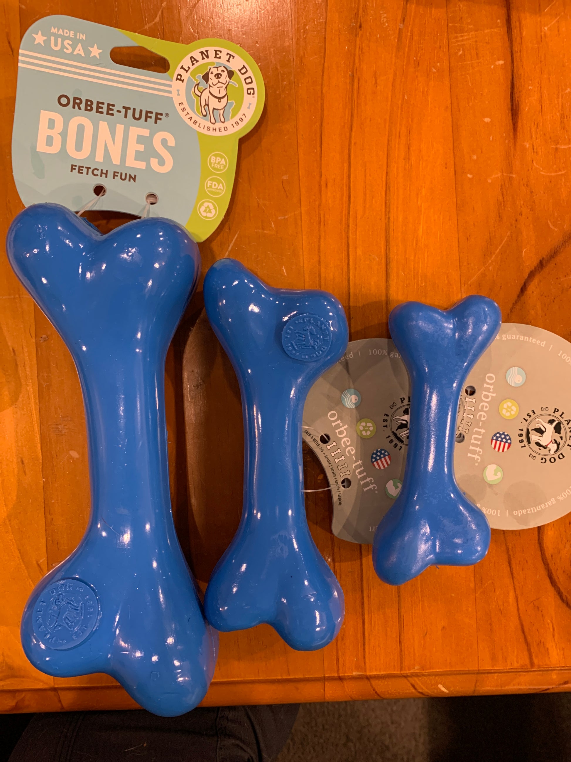 Three blue CLEARANCE: Planet Dog ORBEE-TUFF BONE products of different sizes are displayed on a wooden surface, with tags indicating they are made in the USA by Your Whole Dog, perfect for moderate chewers seeking some chewy goodness.