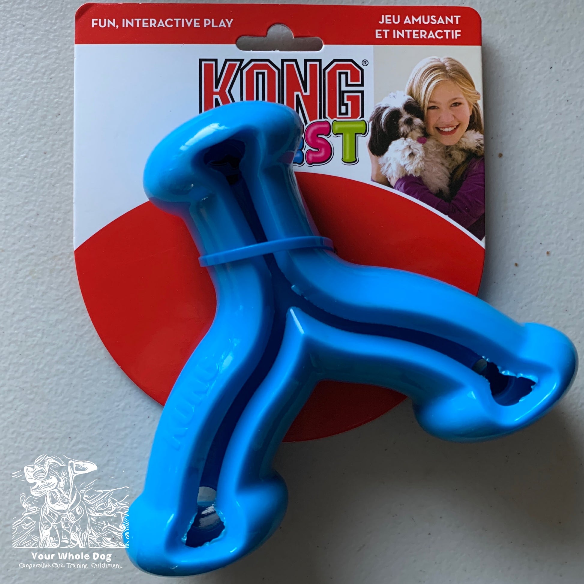 A blue CLEARANCE: KONG Quest Wishbone from Your Whole Dog is attached to its packaging, promoting fun, interactive play in both English and French. The package features a smiling person holding a small dog and highlights the toy's function as a treat-dispensing toy to encourage natural foraging behaviors.