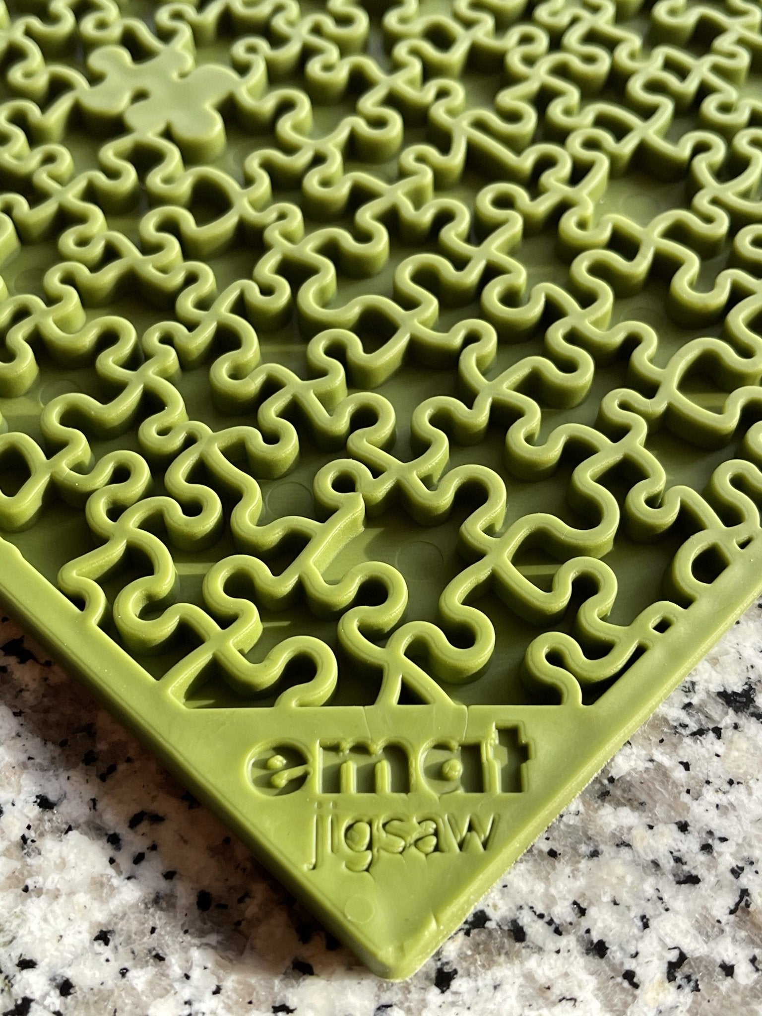 A close-up of a green, Soda Pup EMAT ENRICHMENT LICKING MAT jigsaw puzzle with unique interlocking pieces on a countertop, next to Soda Pup licking enrichment mats.